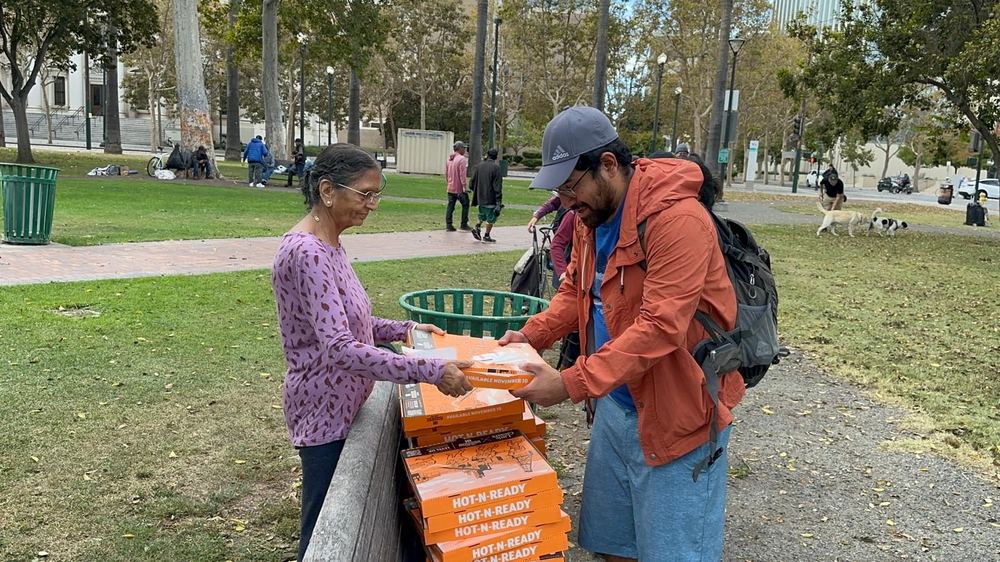 A volunteer hands a boxed meal across the table to a man in the park